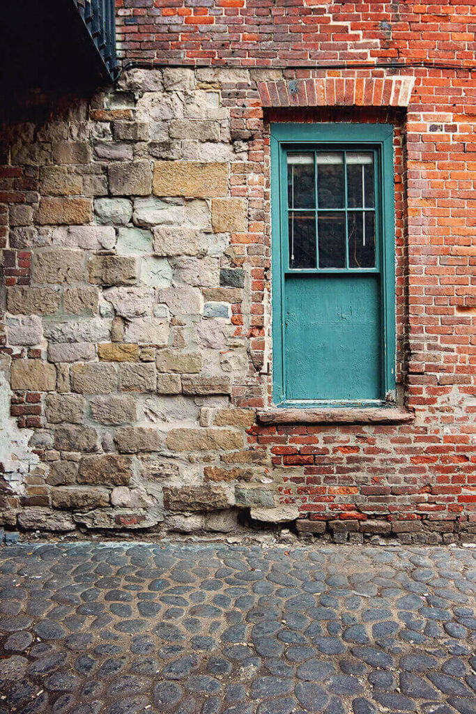 A window frame painted bright teal sits inside an old wall pieced together with various stones and worn red bricks