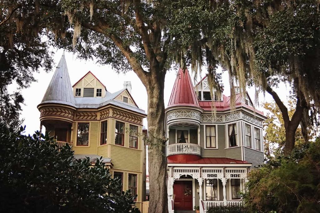 Peering through greenery in the foreground at two "twin" Victorian homes with matching porches and turret-style rooflines