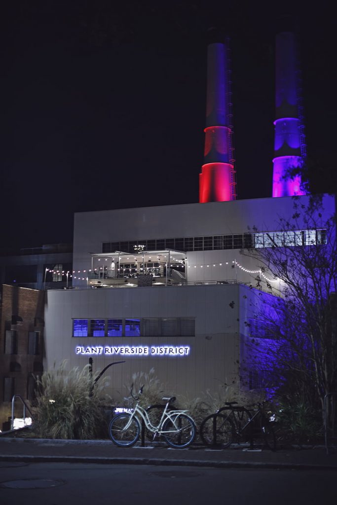 Plant Riverside District sign and smokestacks at night illuminated with purple and red neon lights