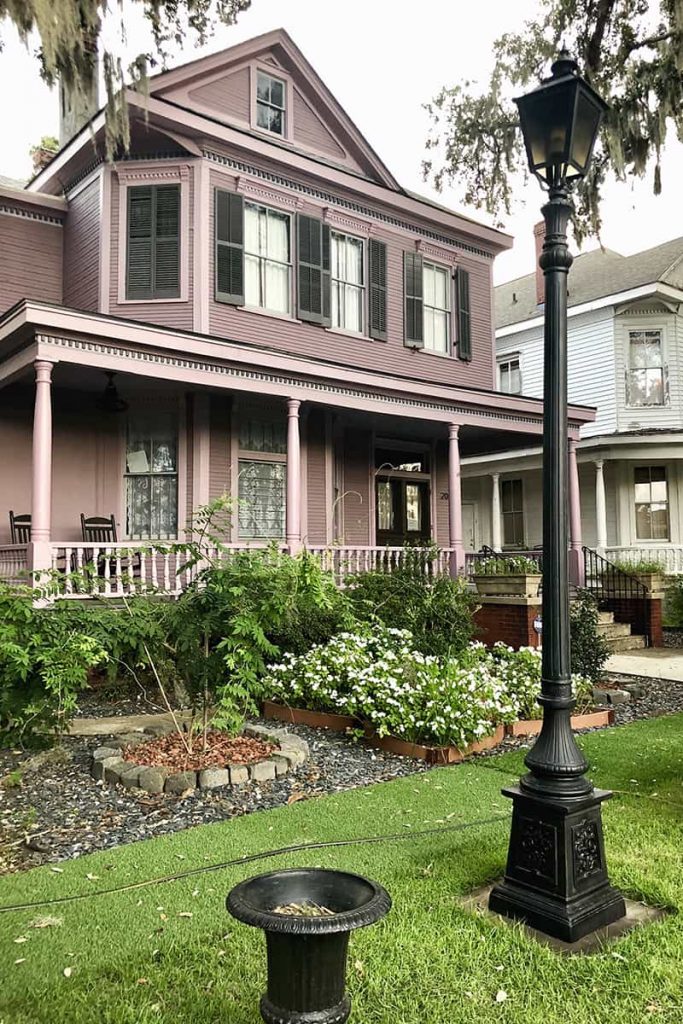 A nicely-landscaped Victorian-style home is painted purple with black shutters. In the foreground it's apparent the sidewalks have been covered with fake bright green grass, and there is an old-timey gas lantern made of cast iron visible in the frame