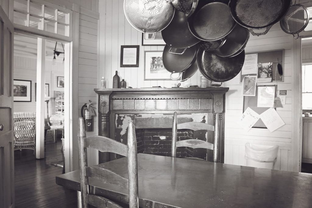 The kitchen of an old cabin shows a wooden table with cast iron post hanging above and a fireplace in the background