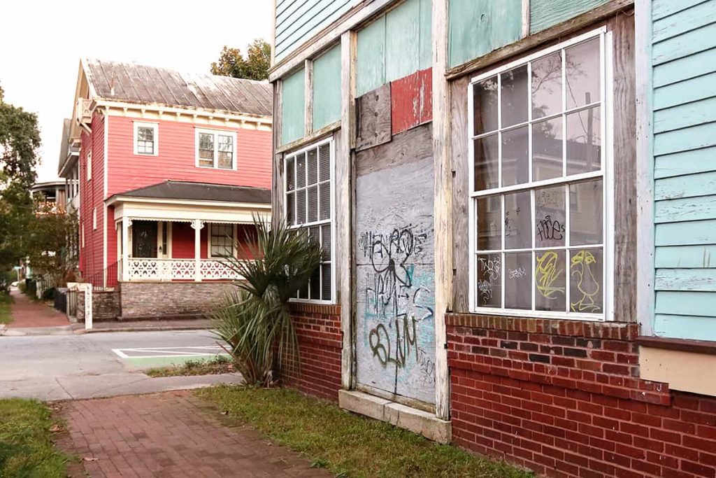 A run-down home with graffiti spray painted on faded plywood boards covering the entrance. A cared-for home can be seen on the corner across the street