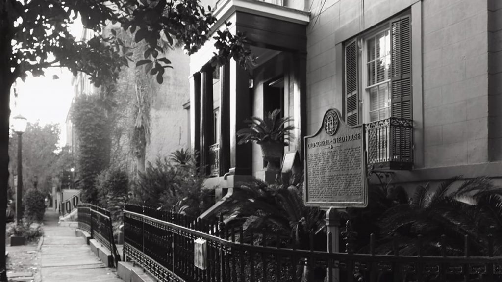 B&W image of the front facade of the Sorrel Weed House in Savannah with a historic marker near the front door and a spiky black wrought-iron fence surrounding the yard