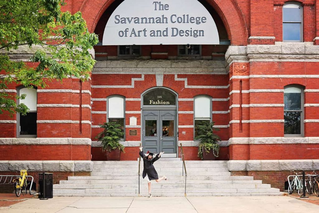 Two-story brick entrance to Eckburg Hall with a large metal sign that reads The Savannah College of Art and Design above the door. A student wearing a graduation cap and gown is leaping in the foreground in front of the staircase