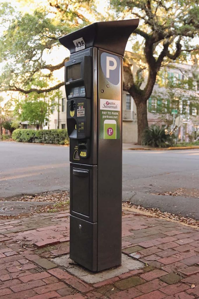An upright parking kiosk on the corner of a street in Savannah with beautiful homes and an old oak tree visible in the background