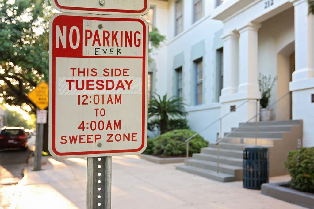 A red and white street sign in Savannah with bold text that reads No Parking (EVER) This Side Tuesday 12:01 AM to 4:00 AM Sweep Zone. The "EVER" was added in black marker by a graffiti artist