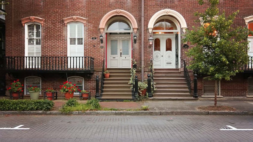 Street scene showing the entrances of two beautiful brick homes with planters filled with red geraniums near the front doors. The road is paved with grey Belgium blocks and white lines with numbers are visible on the road, indicating parking spaces