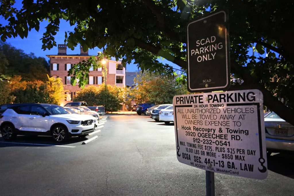 A well-lit and full parking lot at night with a SCAD Parking Only sign and a towing notice for those who break the rules