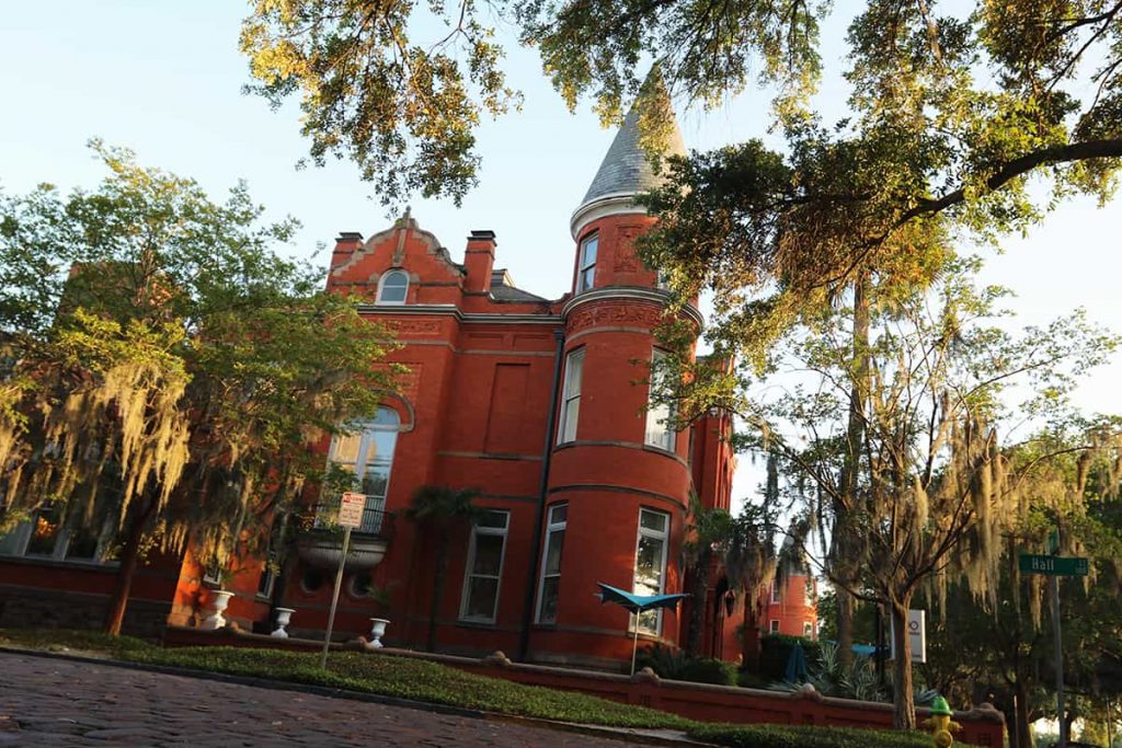 Two-story red brick mansion with a turret on one corner. The photo is shot from a low angle and the sun is setting through the trees, casting a warm glow across the scene