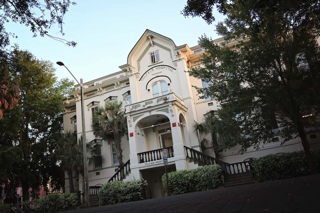 Street view looking up at the front facade of the 3-story white plaster Mary Telfair Hospital. The building is surrounded by trees and the sky is blue in the background