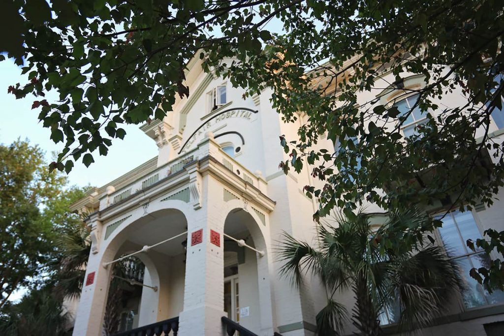 View from the sidewalk looking up through the trees at the imposing entrance of Telfair Hospital in the Victorian District of Savannah. The building is painted in a cream color with sage green trim and has three visible reddish-colored terra cotta tiles in the columns near the entrance