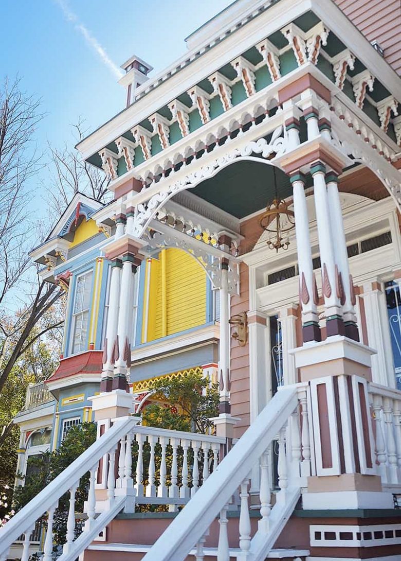 An elaborately trimmed front porch in Savannah's Victorian District is painted pink, green, and burgundy with white trim. A bright yellow and blue Victorian-style home is visible next door
