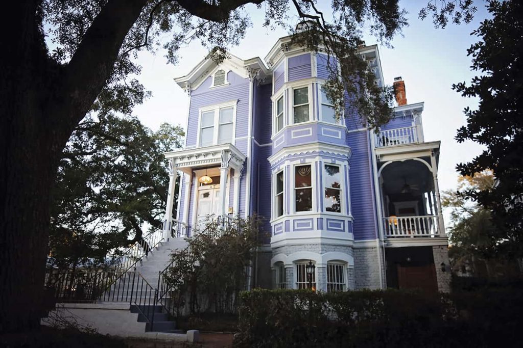 Low-angle view of a 3-story purple Victorian-style home surrounded by trees