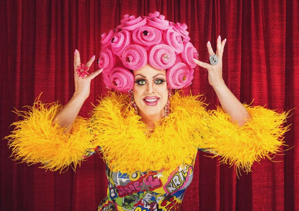 Smiling man dressed as a woman with hot pink cupcake-style curly hair, a feathered yellow top, and a face full of heavy makeup