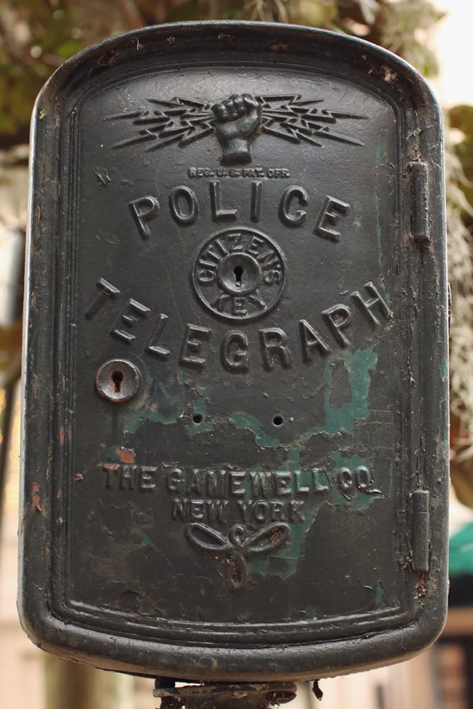 A black wrought-iron police telegraph box in Forsyth Park showing a fist holding lightning bolts and the logo for The Gamewell Co out of New York