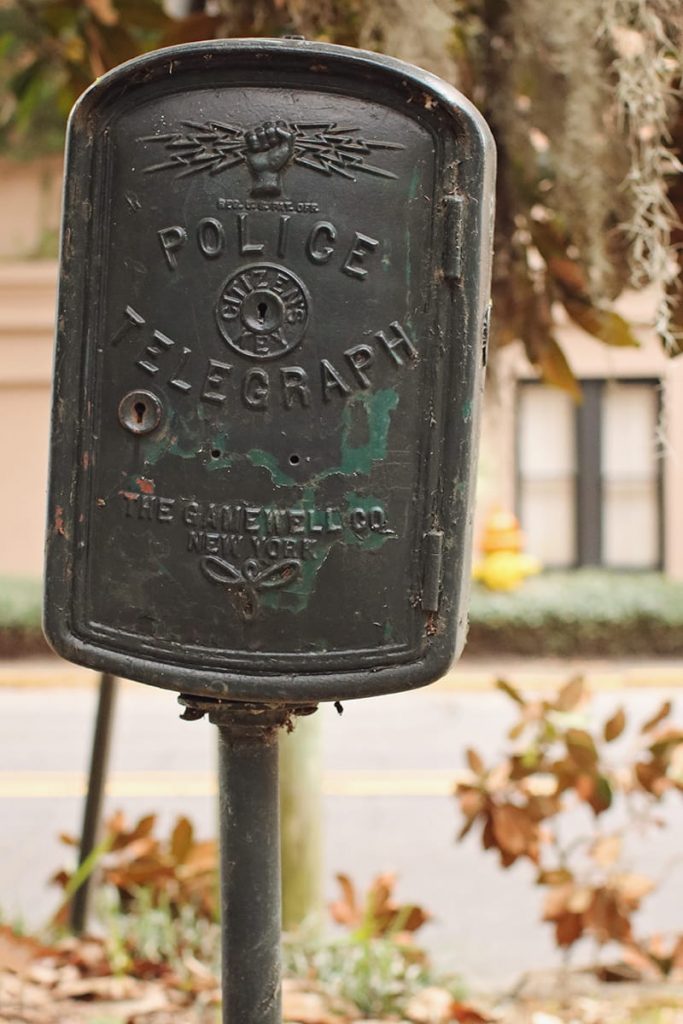 Close-up of a black wrought-iron police telegraph box showing a fist holding lightning bolts and a key insert labeled Citizens Key. Spanish moss and magnolia leaves are visible in the background