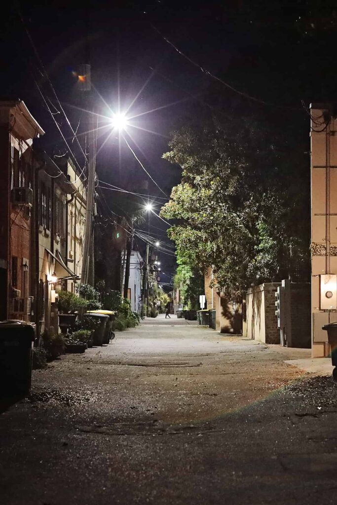 A lane in Savannah with trash cans pushed against garage doors and a bright spotlight illuminating the area. In the distance, the shadow figure of a man walking past the alley is visible