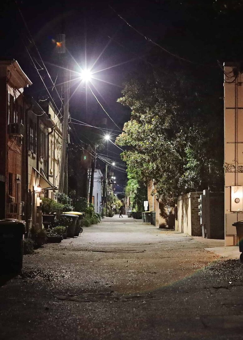 A lane in Savannah with trash cans pushed against garage doors and a bright spotlight illuminating the area. In the distance, the shadow figure of a man walking past the alley is visible