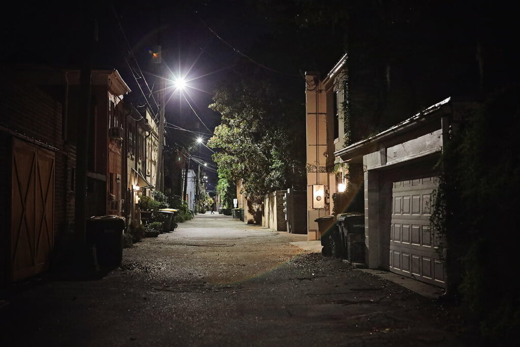 A lane in Savannah with trash cans pushed against garage doors and a bright spotlight illuminating the area. In the distance, the shadow figure of a man walking past the alley is visible