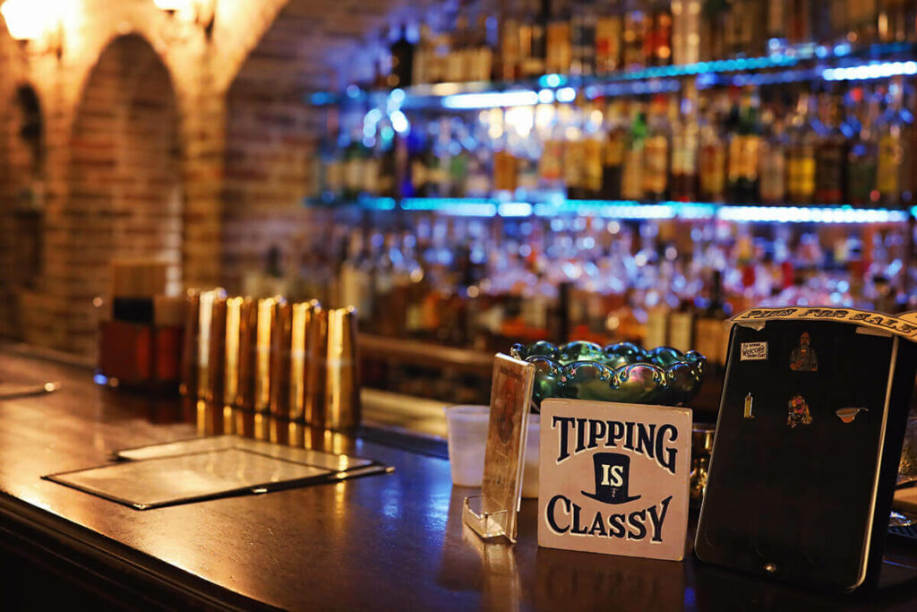 Congress Street Up Speakeasy in Savannah, Georgia, shows a classic-style bar with exposed brick walls, hundreds of liquor bottles, and a sign that reads "tipping is classy"