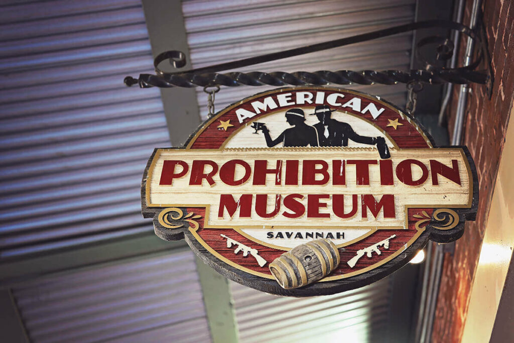 Oval-shaped wooden sign with the words "American Prohibition Museum Savannah" and the image of a flapper-style couple, a barrel of whiskey, and two tommy guns