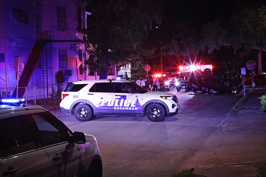A Savannah Police Department vehicle blocks the road where an accident occurred and a car overturned. Blue and red lights illuminate the scene with emergency responders standing amidst broken glass and bystanders