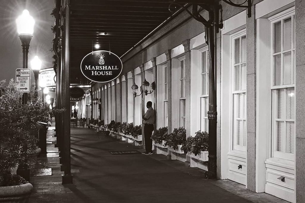 B&W image of the entry level of The Marshall House at night with a doorman standing under a sign with the hotel's logo (a pineapple)