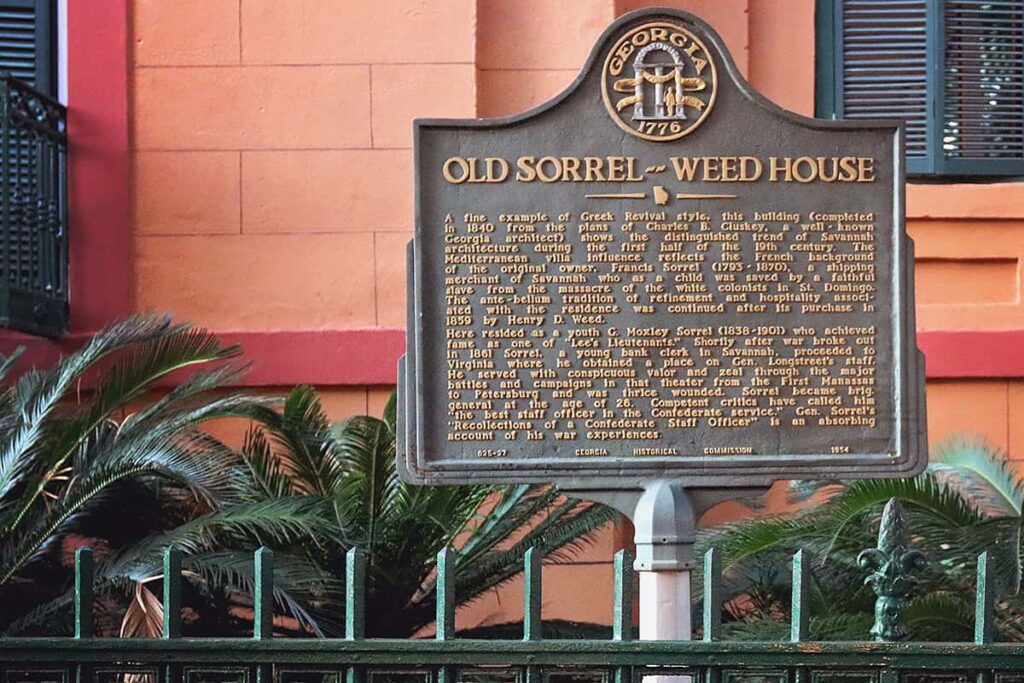 Historic Marker for the Old Sorrel-Weed House with a wrought-iron fence painted green in the foreground and the stucco facade of an orange home with red trim in the background
