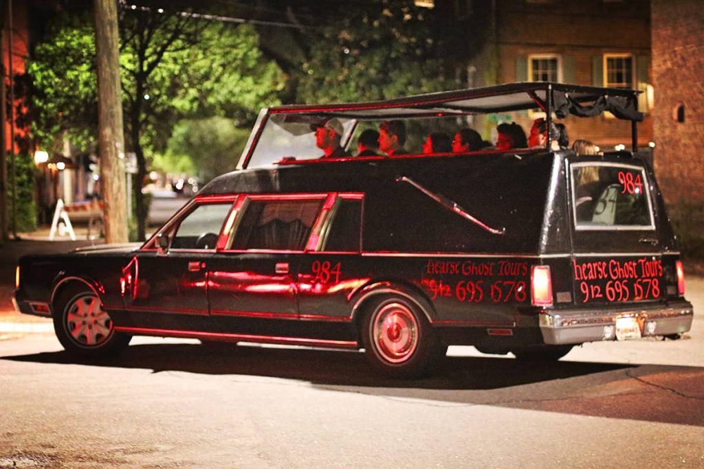 An older model black hearse with red writing on the side drives through Savannah's Historic District with a load of tourists