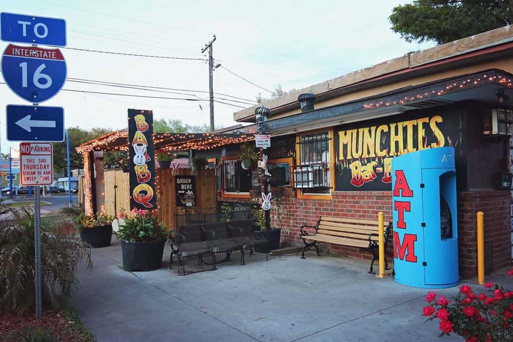 Roadside BBQ stand with a blue interstate sign pointing "this way to HWY 16". The restaurant has festive twinkle lights and a black sign with yellow and red writing that reads "Munchies BBQ" and is made to resemble fire