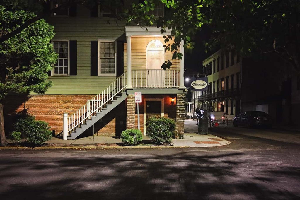 The wood and brick 3-story facade of the 17Hundred90 Inn at night, with spooky shadows from the trees visible on the road in front of the inn. A horse and carriage is traveling on the road beside the inn.