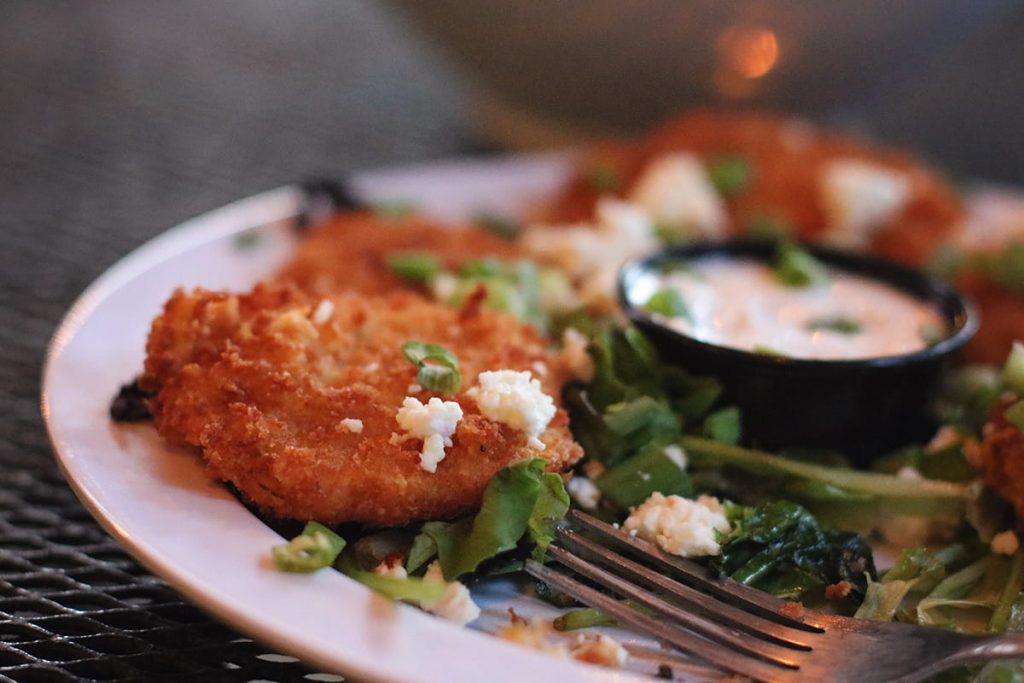 A white plate with crispy fried green tomatoes atop a bed of greens. Goat cheese crumbles are sprinkled atop the tomatoes and a side dish of Buttermilk dressing is in the center of the plate