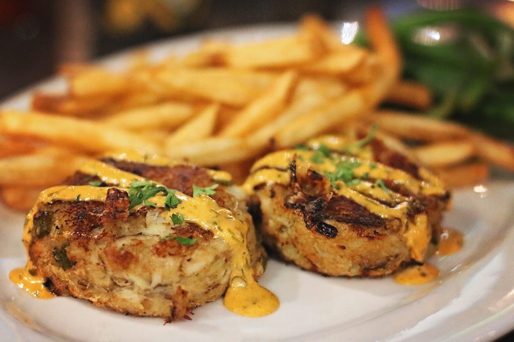Two golden fried crab cakes drizzled with a tangy yellow sauce and bits of green spices. Crispy golden fries and green beans can be seen in the background of the plate