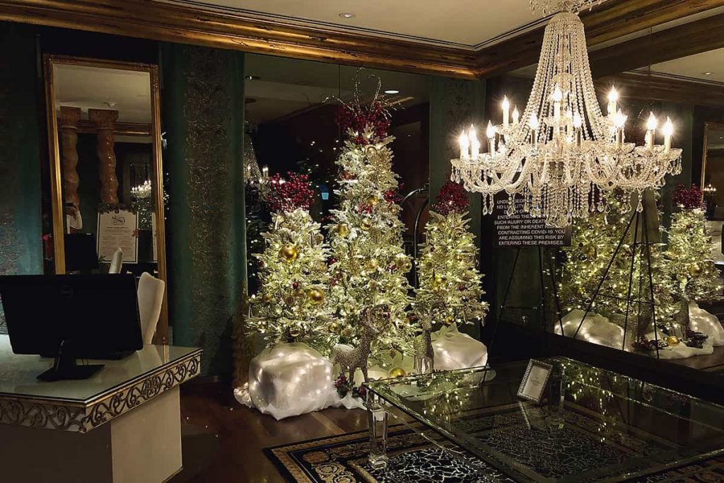 Opulent lobby of The Mansion on Forsyth showing three Christmas trees made of feathers and decorated with gold globes and red berries. A sparkly crystal chandelier hangs from the ceiling, adding a warm glow to the gilded trim along the ceiling