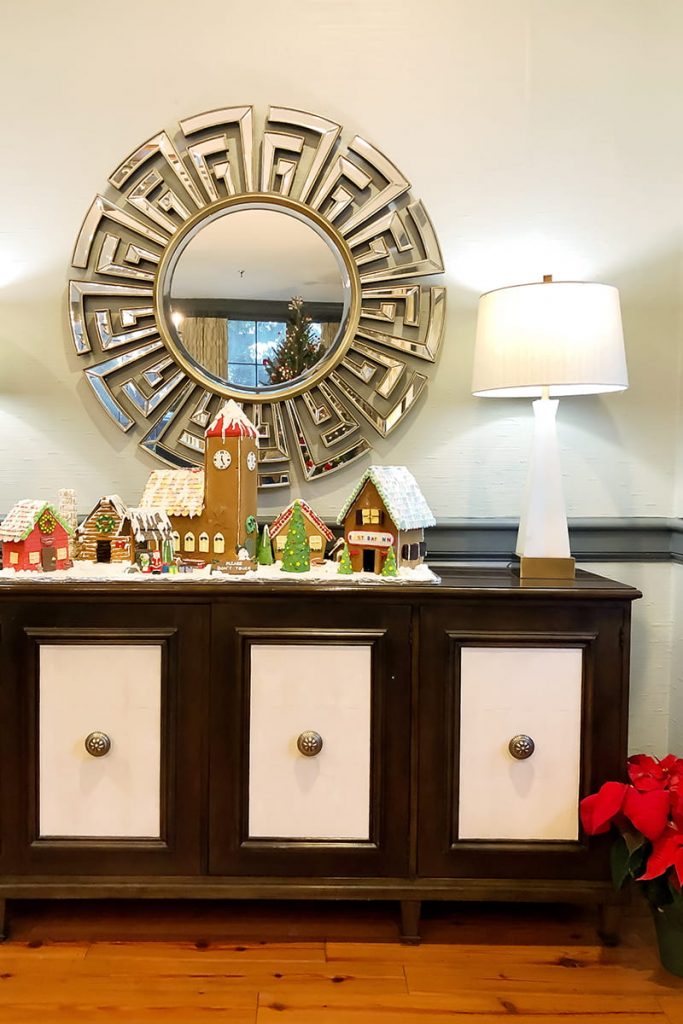 Christmas gingerbread village set up on a brown and white credenza in the lobby of East Bay Inn. A Christmas tree can be seen reflected in the mirror behind the village