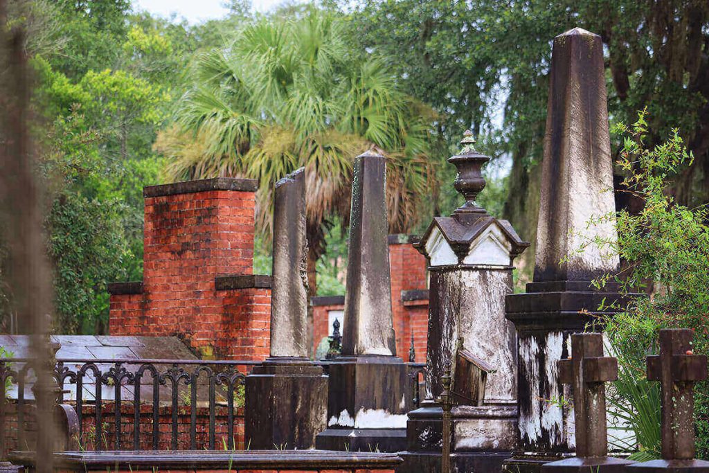 Obelisk-style monuments in Laurel Grove Cemetery with dark brown coloring indicating their age. A brick vault and lush greenery can be seen in the background