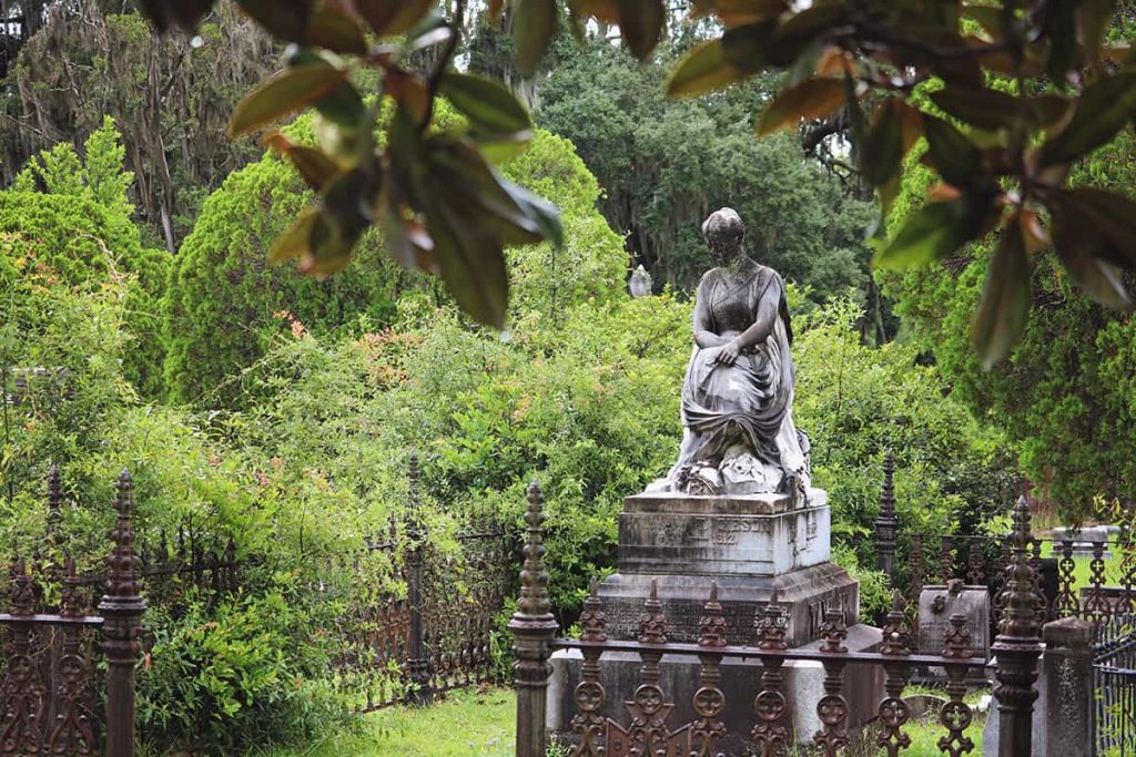 Peering through Magnolia leaves at a statue in Laurel Grove Cemetery of a woman seated with her arms and legs crossed. Very lush greenery can be seen in the background, and the plot is surrounded by intricate wrought-iron fencing