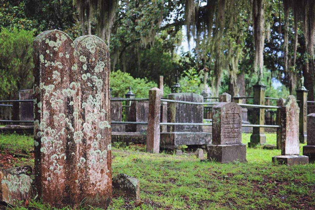 Two tall twin headstones covered in moss and lichens in the foreground with headstones and old oaks in the background