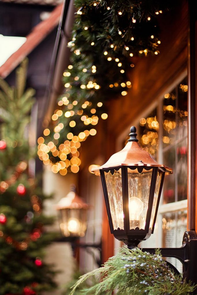 Exterior of an inn decorated for Christmas with greenery and twinkle lights visible behind an old-timey gas lantern