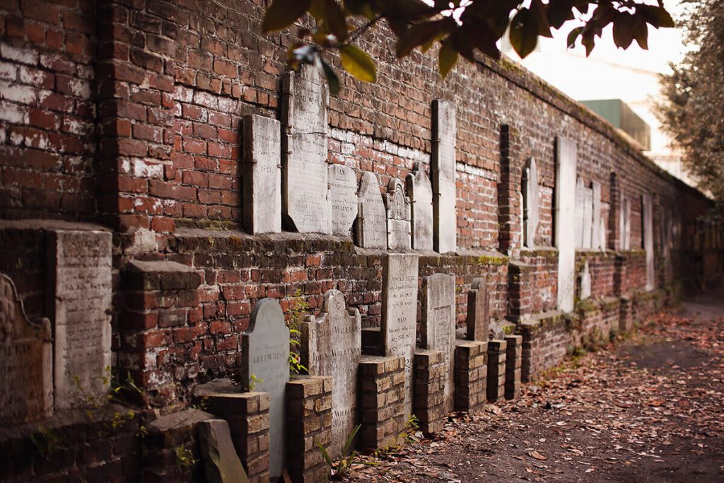 An old brick wall in Colonial Park Cemetery with old headstones affixed to it. Fall leaves are scattered in the foreground