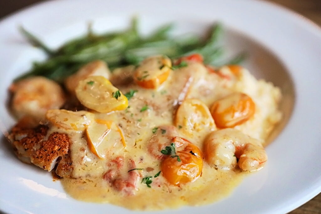 Pan-seared redfish, heirloom tomatoes, and shrimp over grits in a creamy garlic sauce with green beans visible in the background