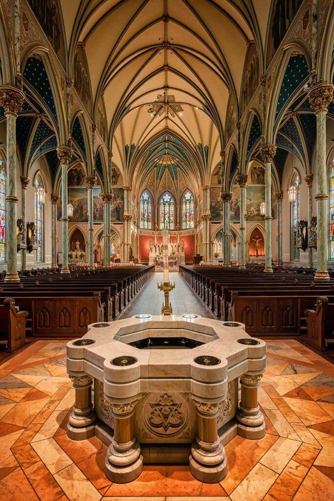A heavy marble baptismal font inside the Cathedral of St. John the Baptist surrounded by decorative floor tiles. The alter is visible in the background with rows of approximately 20 pews leading towards it