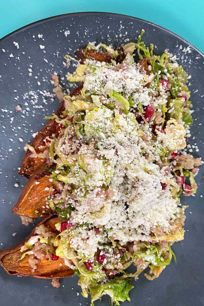 Slate grey colored plate with baked sweet potatoes sliced open and covered with field greens, radishes, and a heavy sprinkling of parmesan cheese