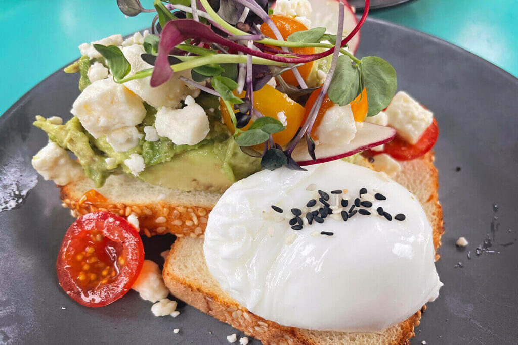 Grey plate from The Colllins Quarter with two slices of bread topped with eggs, avocado, fresh tomatoes, and other colorful veggies