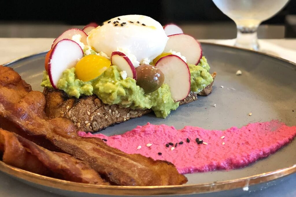 Grey plate with three slices of crisp bacon, and a slice of wheat toast covered with avocado, beets, heirloom tomatoes, and an egg. The plate is decorated with a swipe of mashed red beet hummus sprinkled in seeds