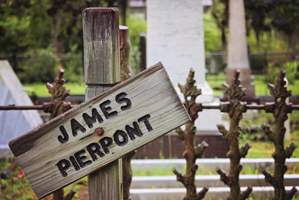 A crooked wooden sign with the words James Pierpont etched into the surface. Rusted wrought-iron spikes are visible behind it, with thick headstones in the background