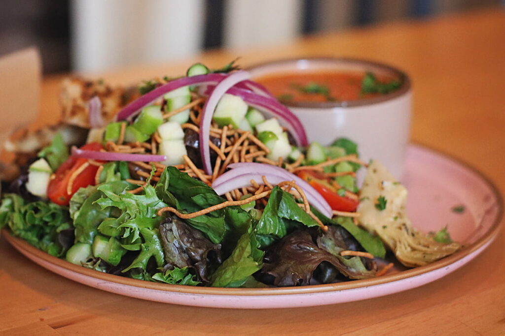 Plate loaded with a salad of dark green lettuce leaves, tomatoes, green apples, and red onion. A bowl of tomato soup is visible in the background