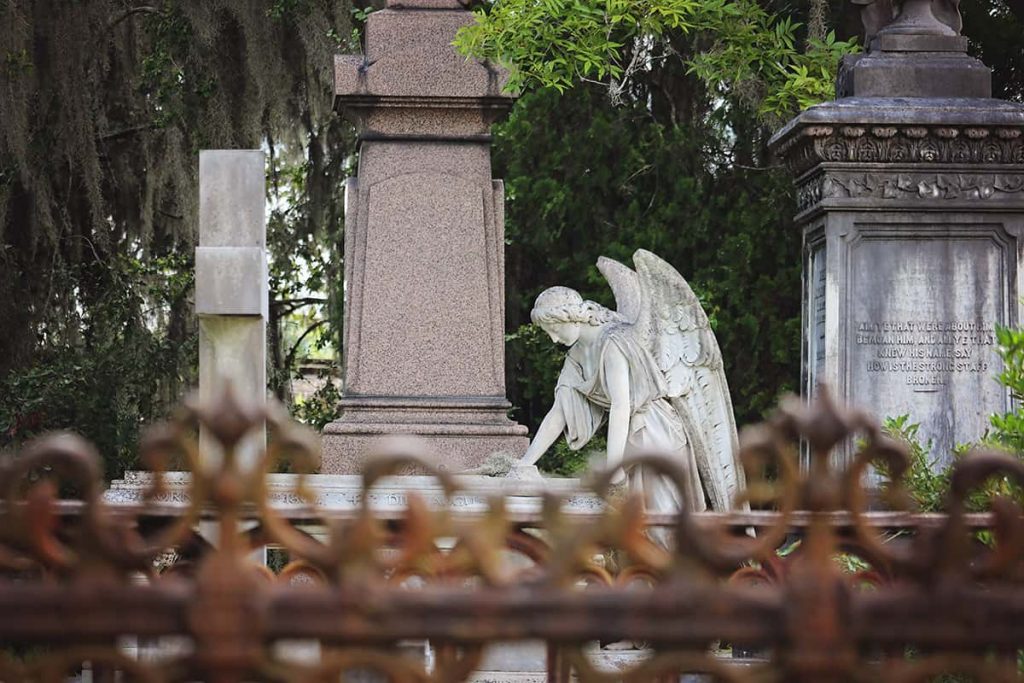 An intricate rusted wrought-iron fence surrounds a cemetery plot in the foreground, while four elaborate Victorian-era statues are visible amongst a backdrop of oaks and Spanish moss in the background of Laurel Grove Cemetery