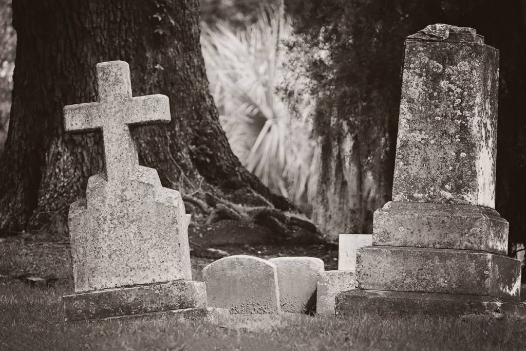 A heavy cross marker on the left and a solid stone marker on the right, both slightly askew from ground settling in Laurel Grove Cemetery. Massive oaks and Spanish moss can be seen in the background