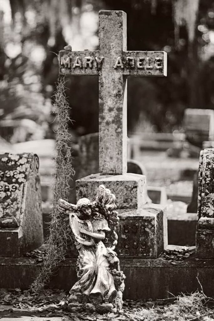 B&W shot of a simple marble cross in a cemetery with the name Mary Adele in raised lettering. Spanish moss hangs from the cross and a small angel statue has been placed in front of the marker